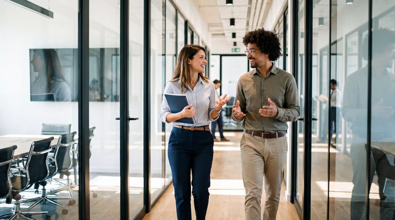 Two colleagues talking as they walk along a glass-walled corridor